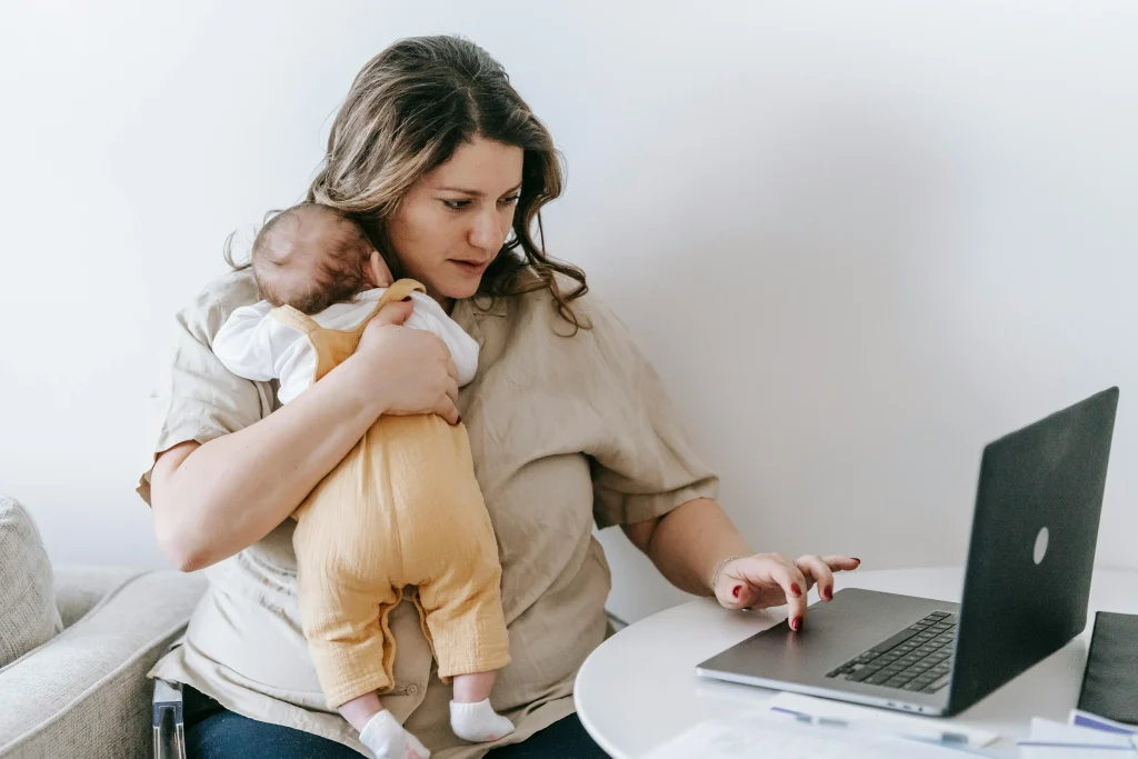Mère portant son bébé tout en travaillant sur son ordinateur, illustrant les enjeux de conciliation entre parentalité et santé mentale au travail.